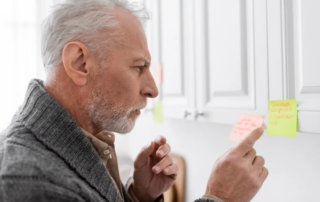 Thoughtful man with Alzheimer’s syndrome pointing at blurred sticky note in kitchen.